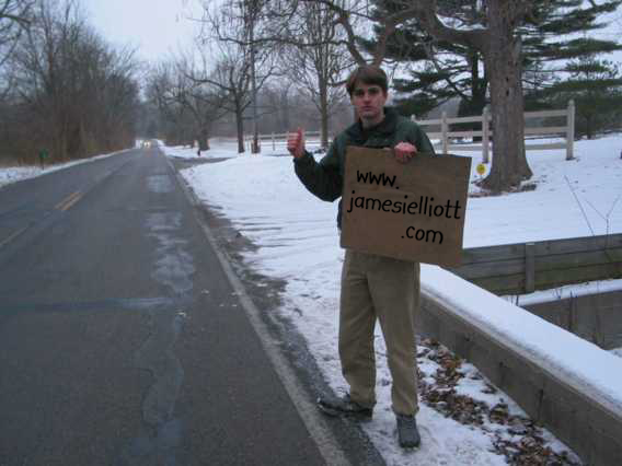 A low-resolution photo of a white high school boy in a green winter jacket. He is standing at the side of a country road trying to thumb a ride while holding a piece of cardboard that reads w w w dot james i elliott dot com. There's snow on the ground and bare trees in the background.